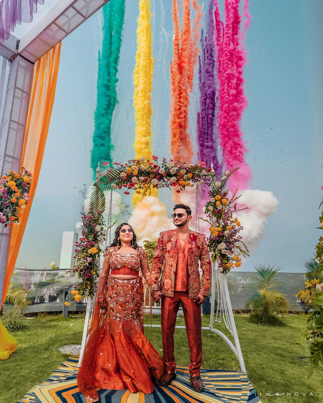 Couple in matching reddish attire holding hands in front of a floral arch as vibrant plumes of green, yellow, and pink smoke rise in the background, captured