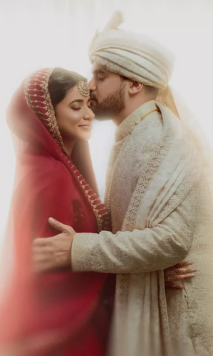 Groom in an embroidered sherwani and turban kisses the forehead of a bride wearing a red hooded veil, photographed by Wedding Photographers in kolkata