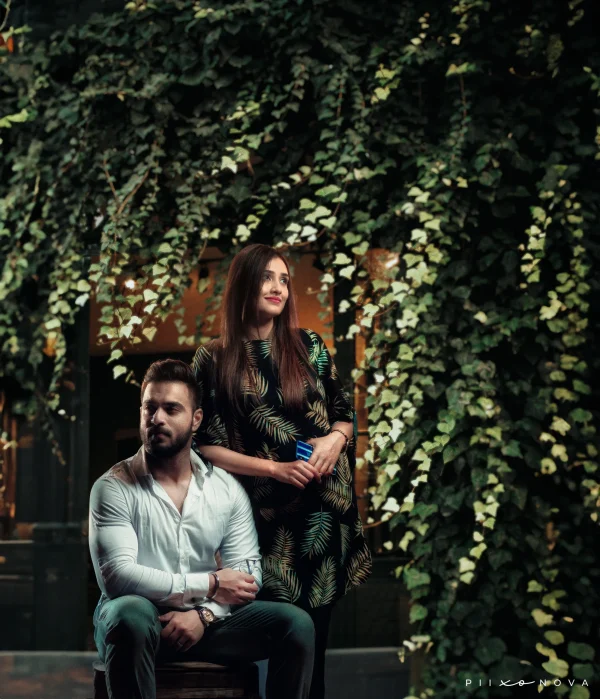 A couple posing together in a lush, green setting. The man sits on a stool, wearing a white shirt and green pants, while the woman stands beside him in a patterned dress, smiling softly against a backdrop of vibrant foliage. Shot by Piixonova