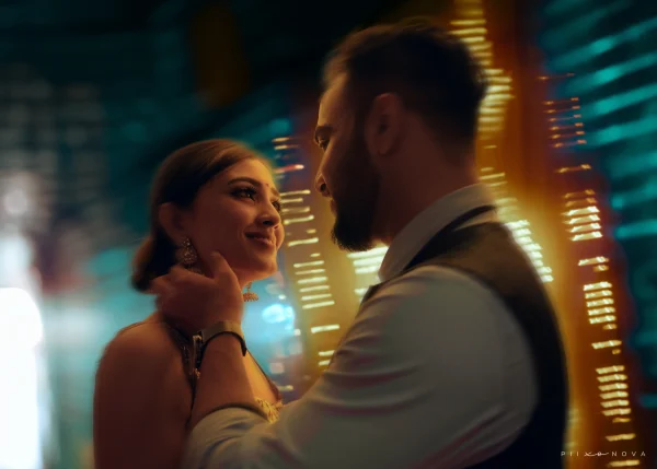 A romantic moment between a couple, with the woman smiling softly as the man gently touches her face. The background features vibrant, blurred lights, creating a dreamy atmosphere. Shot by Piixonova