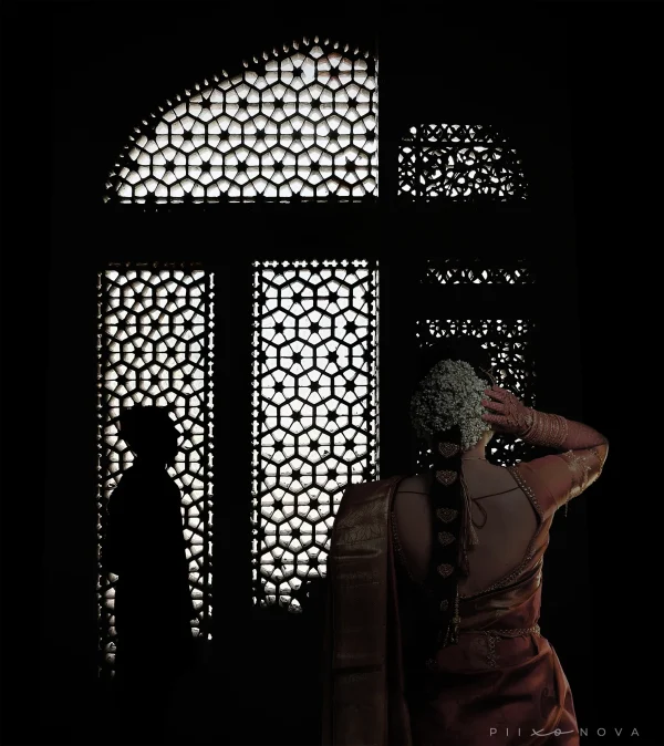 A woman stands gracefully in front of a beautifully designed window, showcasing intricate patterns and details and the groom silhouette can be seen in the distance. Shot by Piixonova